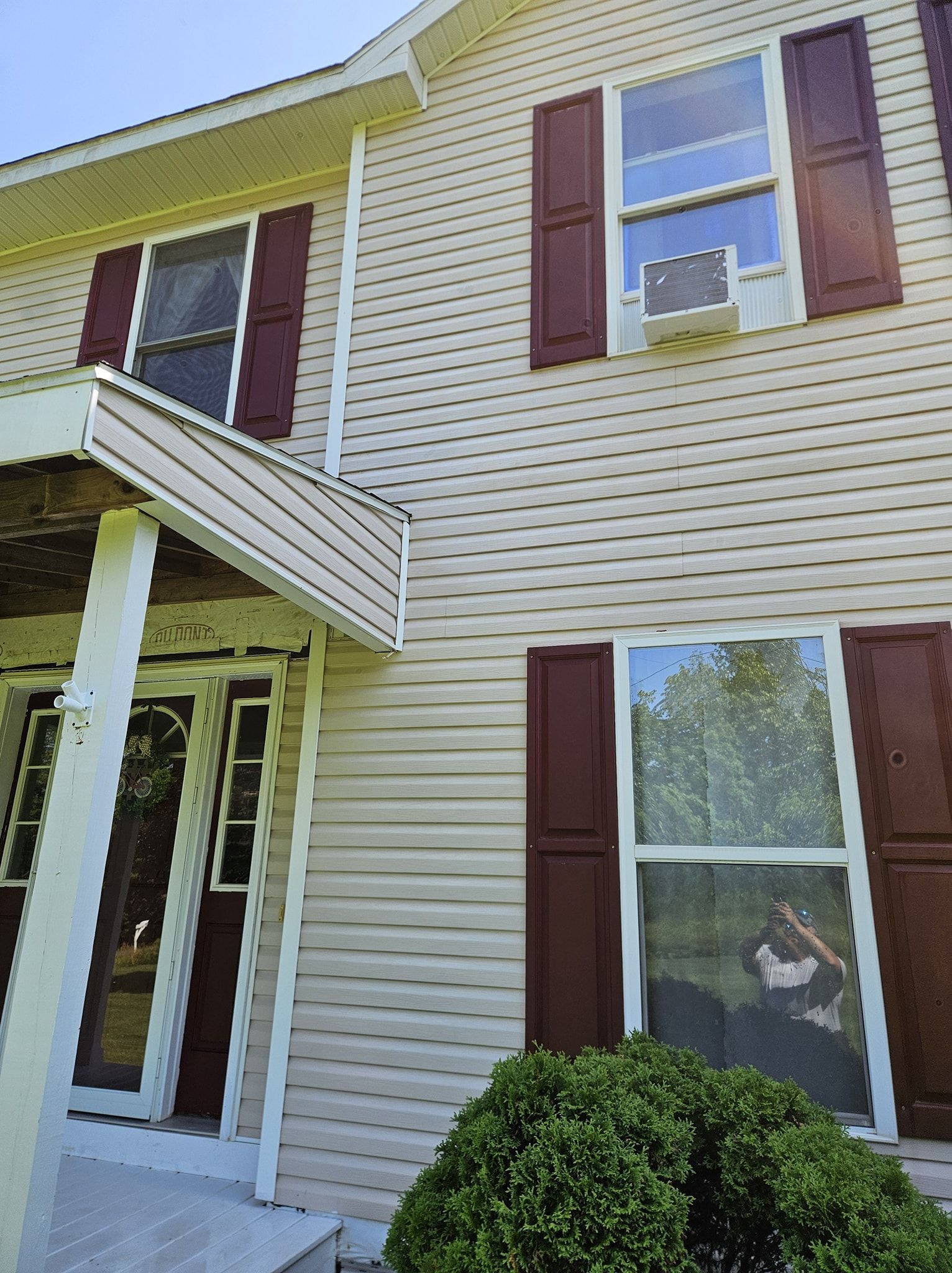 Beige vinyl-sided two-story house exterior with a small front porch, dark red shutters, and a window air conditioner.