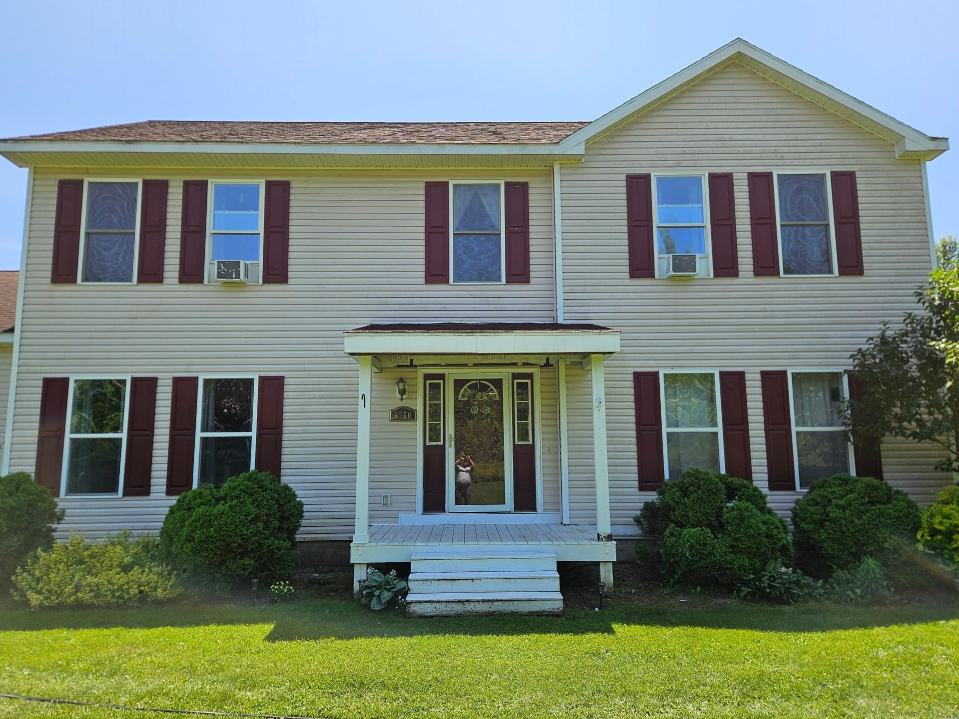 A two-story, light-colored suburban house with red shutters, a front porch, and trimmed green bushes in the front yard.