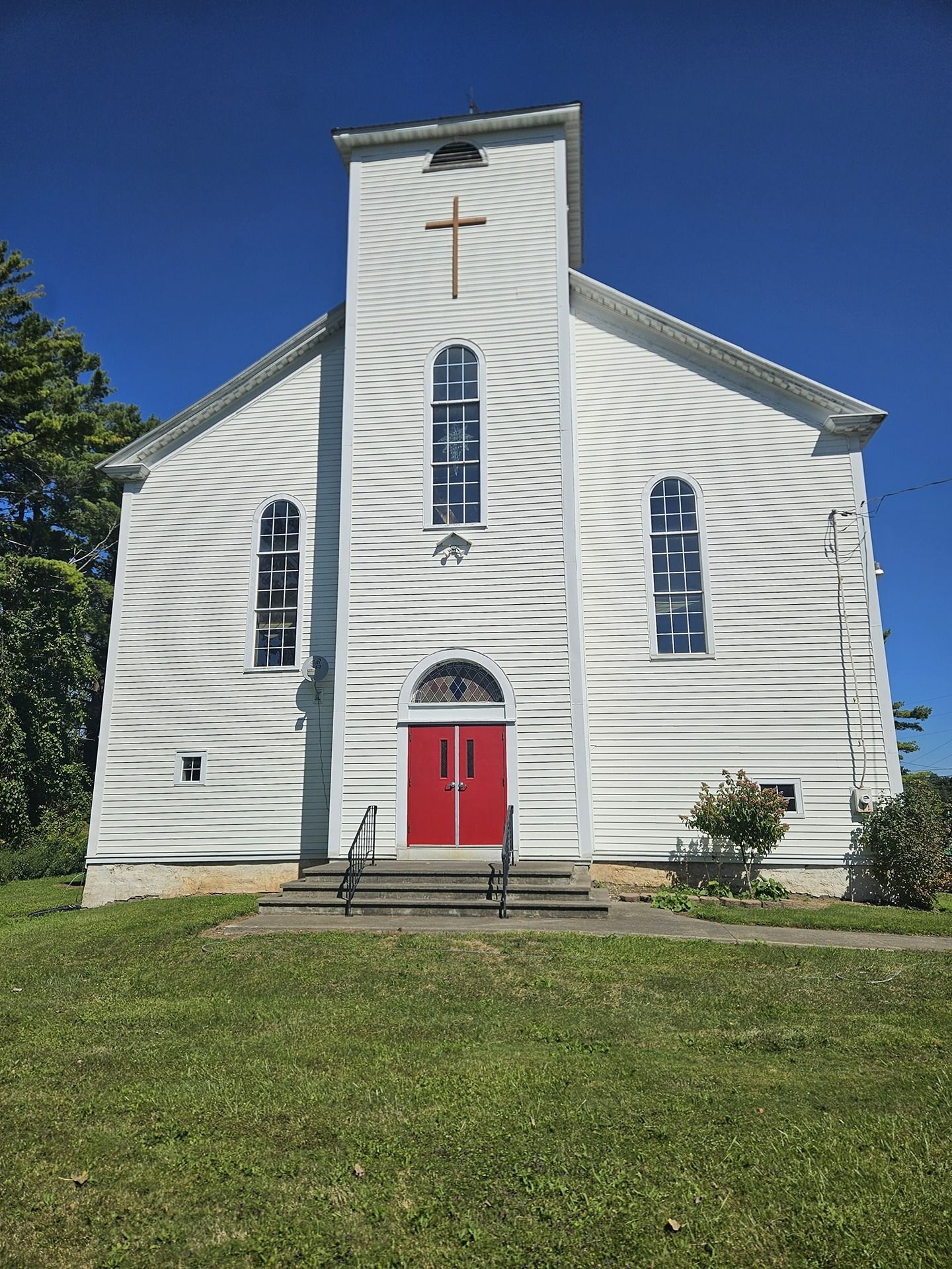 A white clapboard church with a central tower, a prominent red double door, and arched windows under a bright blue sky.