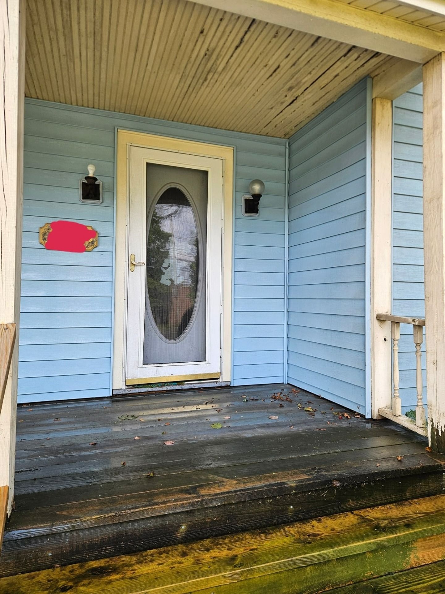 A light blue house front porch with a white door featuring an oval glass window, two wall lamps, and a wooden floor.
