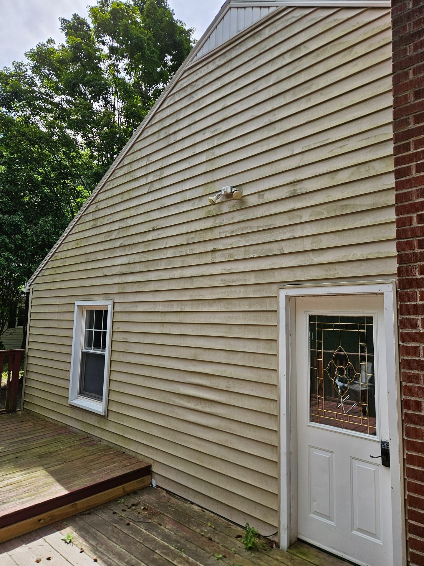 The side of a house with light tan horizontal siding, a white window, a white door with glass panels, and a wood deck.
