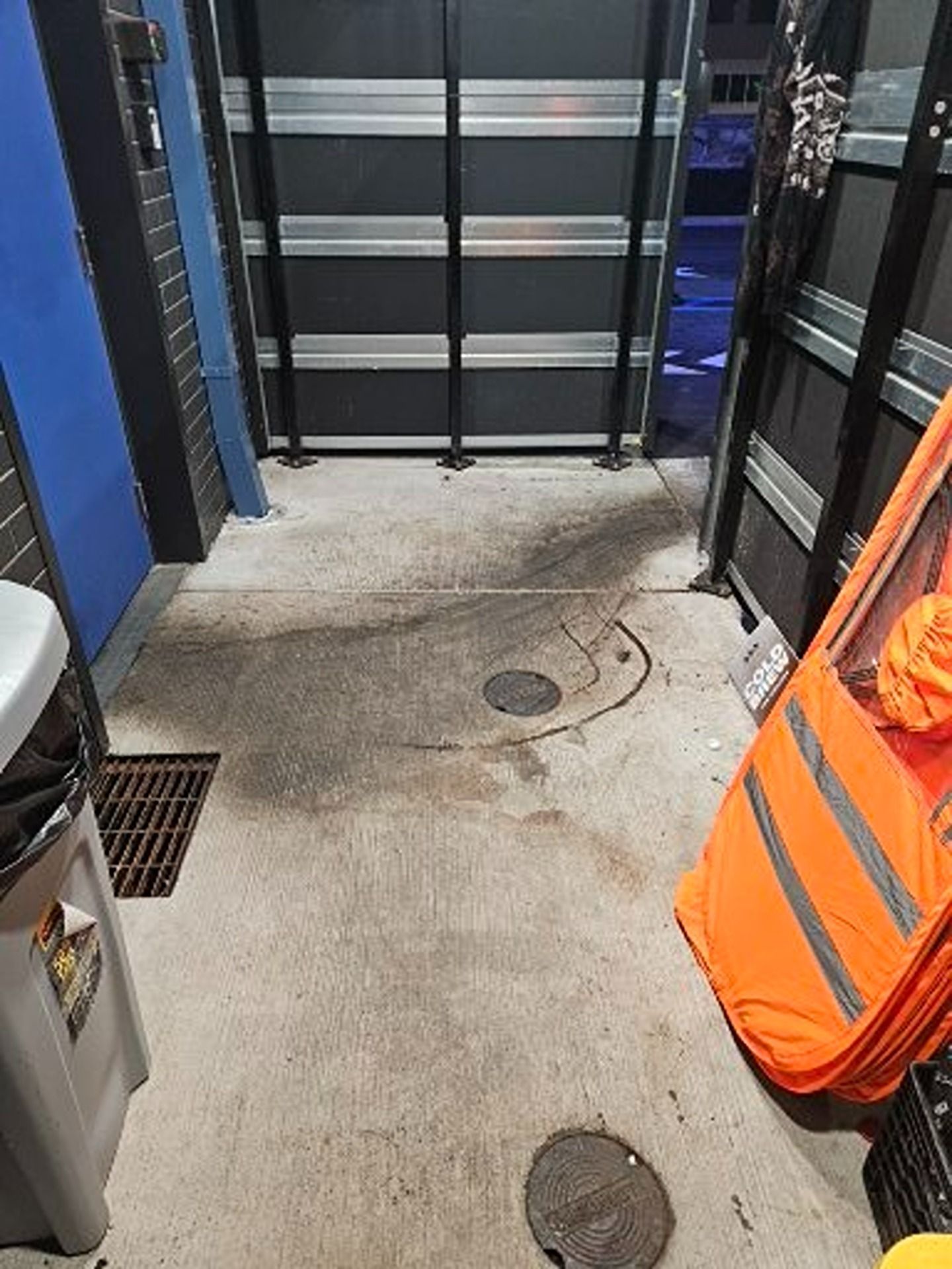 A concrete floor leading to a dark rolling garage door, featuring a drainage grate, a manhole cover, and an orange screen.