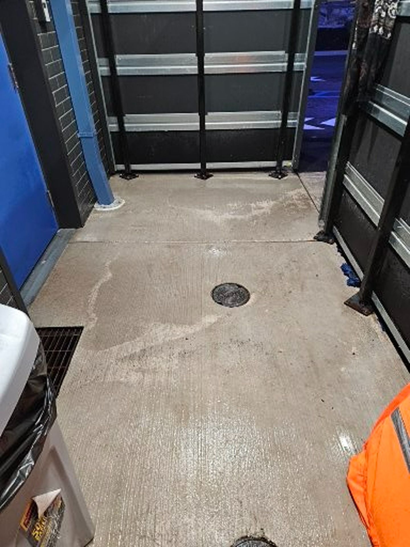 A concrete floor in a workspace with a round floor drain, a blue wall on the left, metal shelving, and a trash bin.
