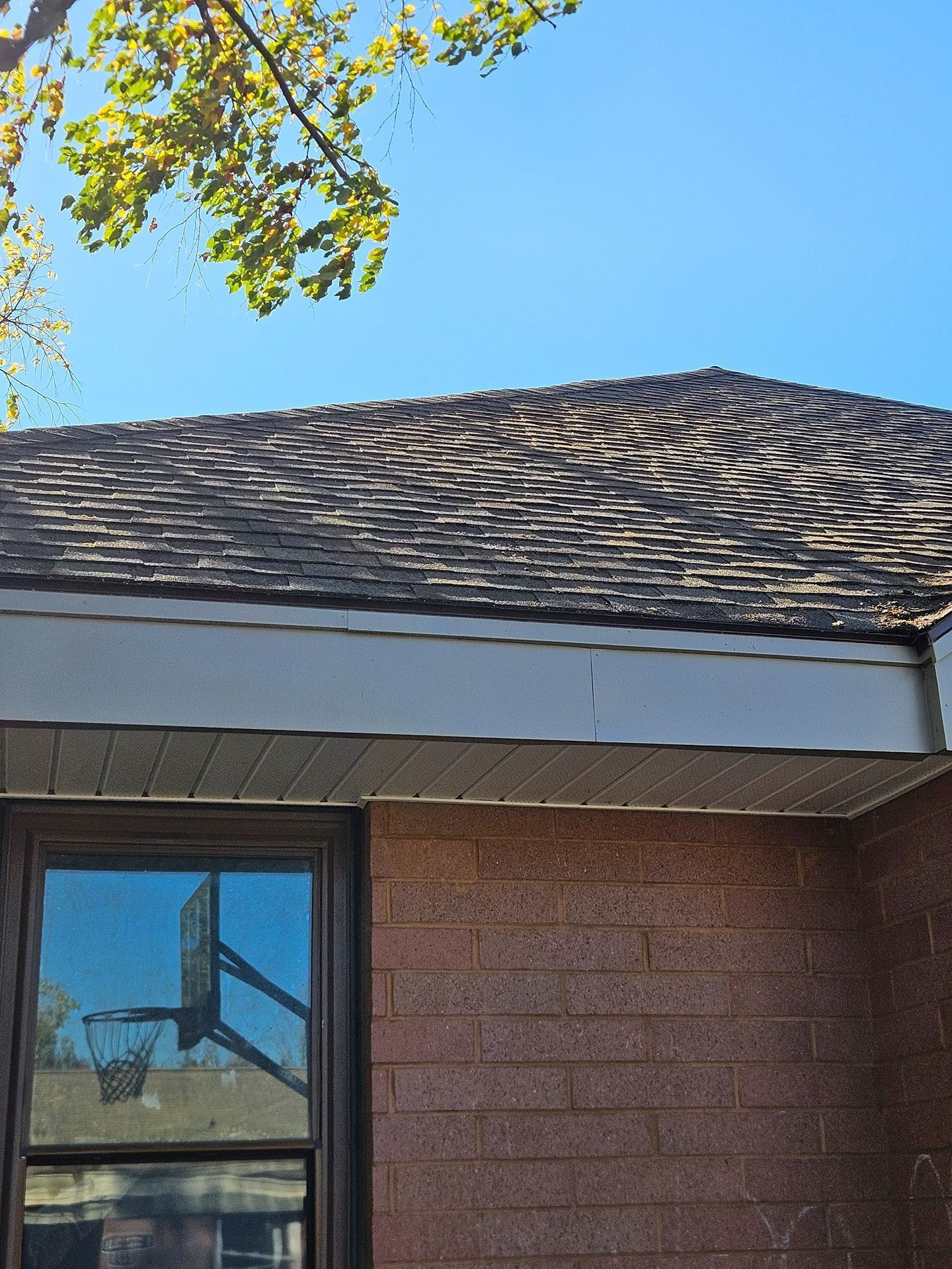 A brick house exterior with a dark shingled roof and a window reflecting a basketball hoop under a clear blue sky.