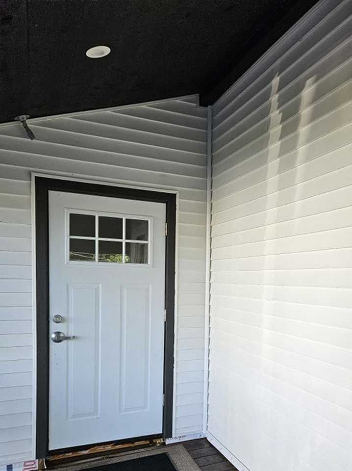 A white door with a six-pane window sits under a porch, next to light gray siding with visible vertical water streaks.