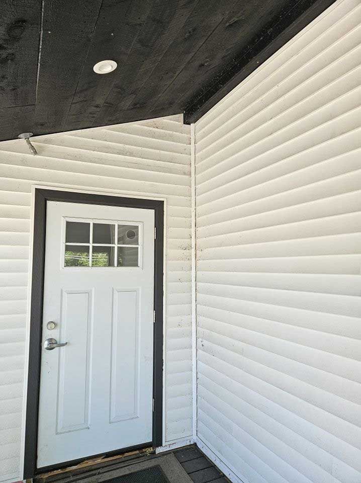 A white door with a window sits on a porch with white siding and a dark ceiling.