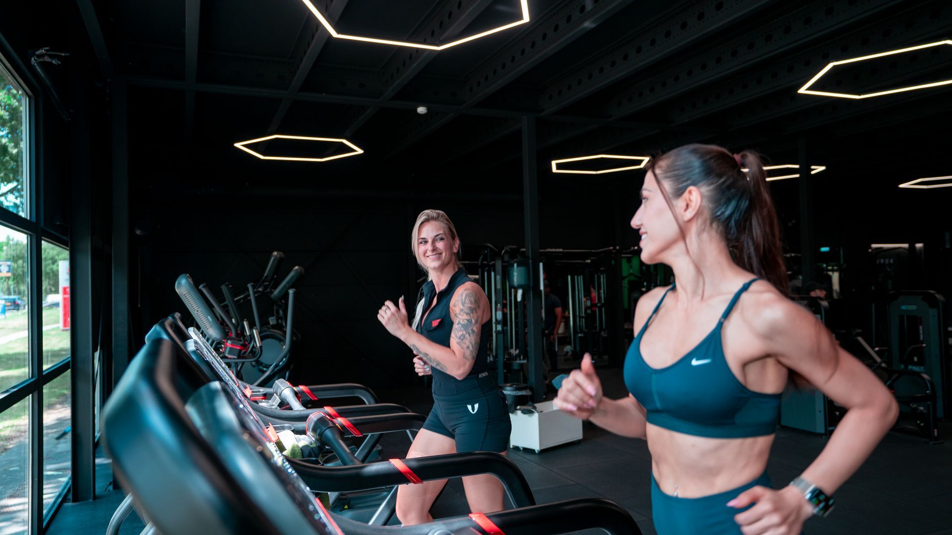 Twee vrouwen in sport-bh's glimlachen op loopbanden in een gedimd verlichte sportschool.