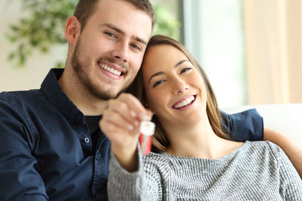 A Man And A Woman Are Sitting On A Couch Holding Keys To Their New Home — Settle Petal Mobile Conveyancing In Wallabi Point, NSW