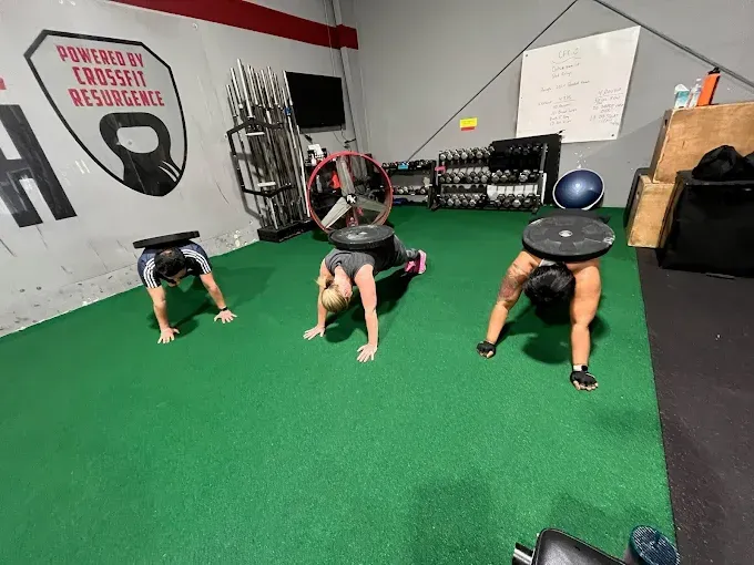Three people in Resurgence Strength in Naperville, IL holding a plank position on green turf with exercise equipment in the background