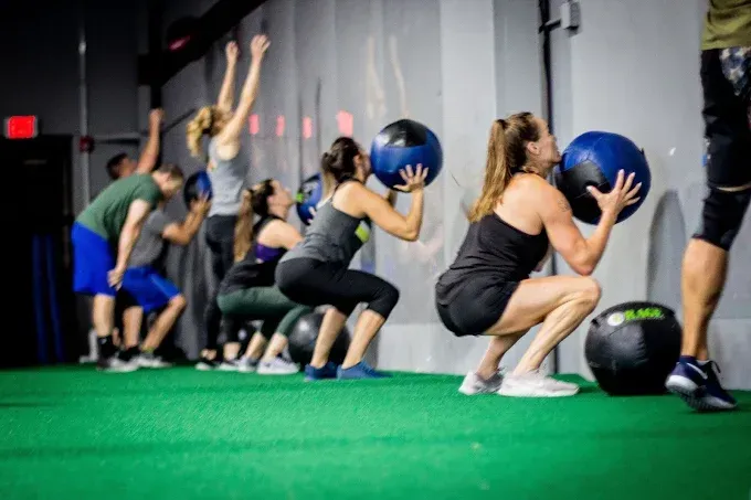 People exercising squat holds with medicine balls in a gym, on green turf in front of a mirror at Resurgence Strength in Naperville, IL