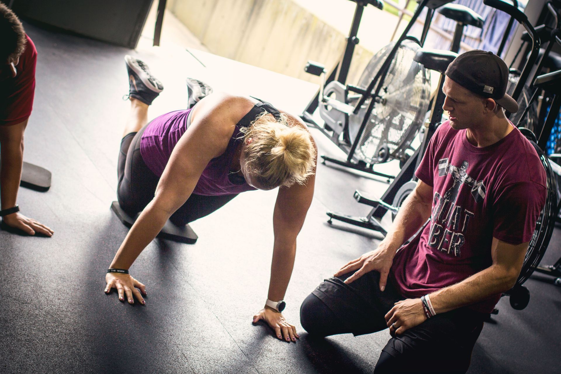 Two people in a gym adjusting a black exercise machine near hanging punching bags.