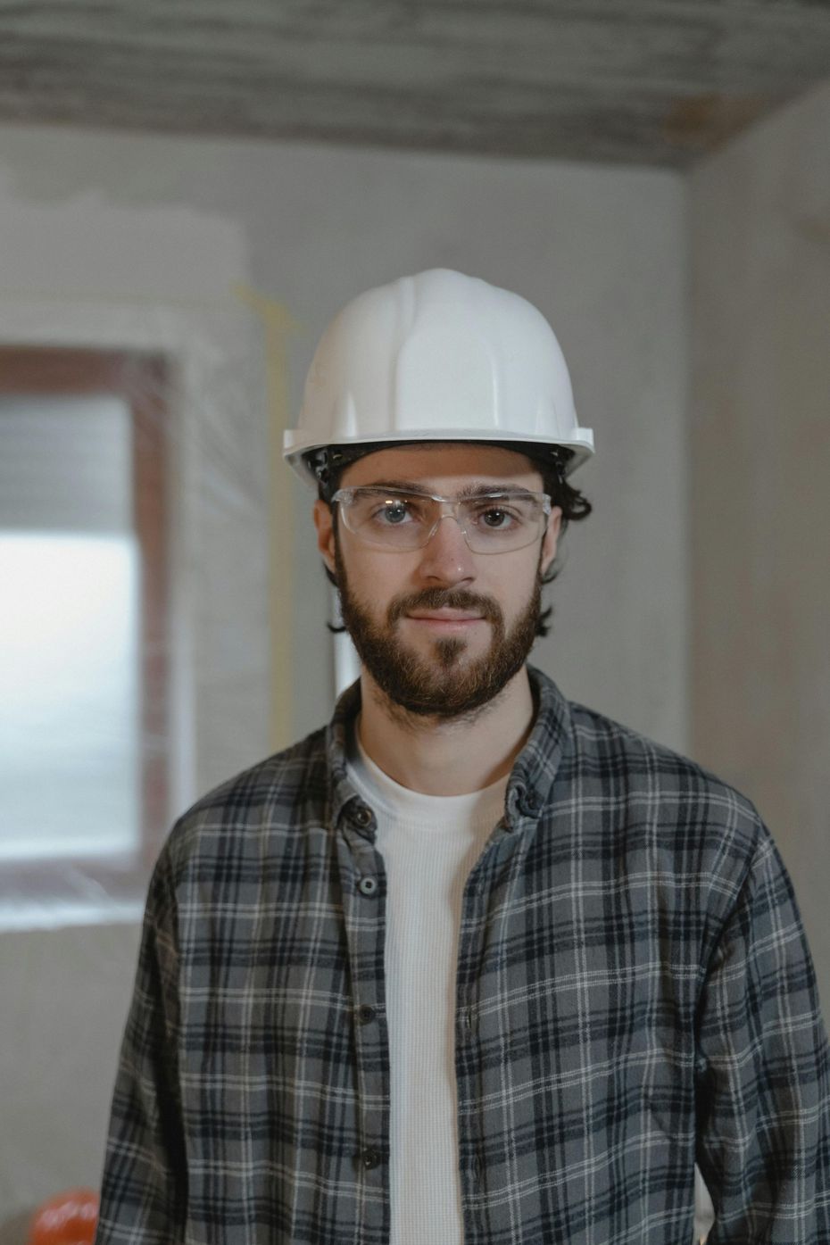 Man in a hard hat and safety glasses, wearing a plaid shirt. Inside a building under construction.