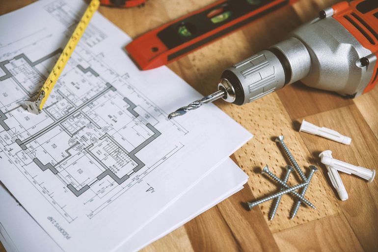 Blueprint with drill, level, tape measure, nails, and wall anchors on wooden surface.