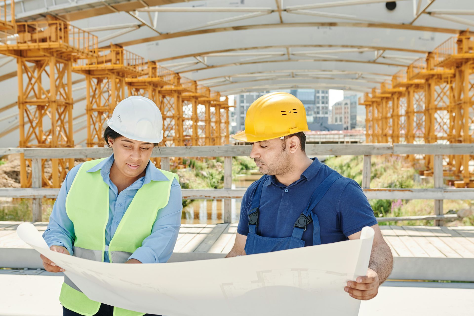 Two construction workers in hard hats examine blueprints at a construction site.