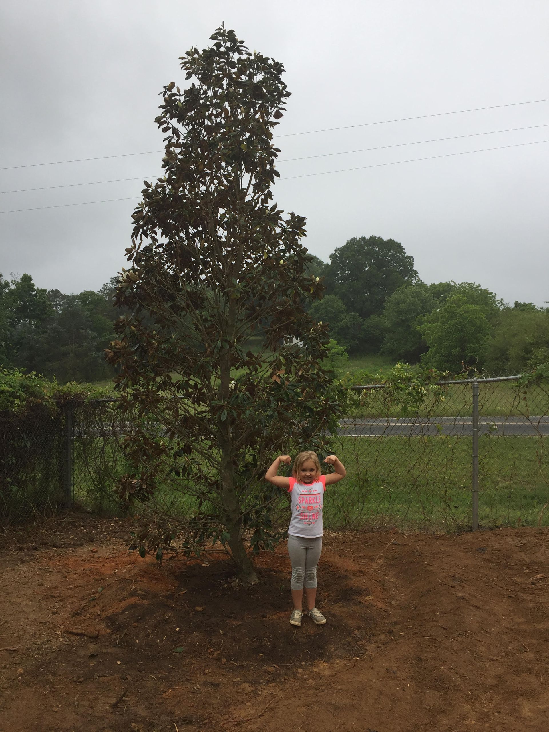 Young girl flexing arms next to a tall tree. Cloudy day, dirt ground, and background trees.