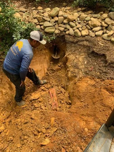 Man inspecting buried pipe in a dirt trench, surrounded by rocks and vegetation.