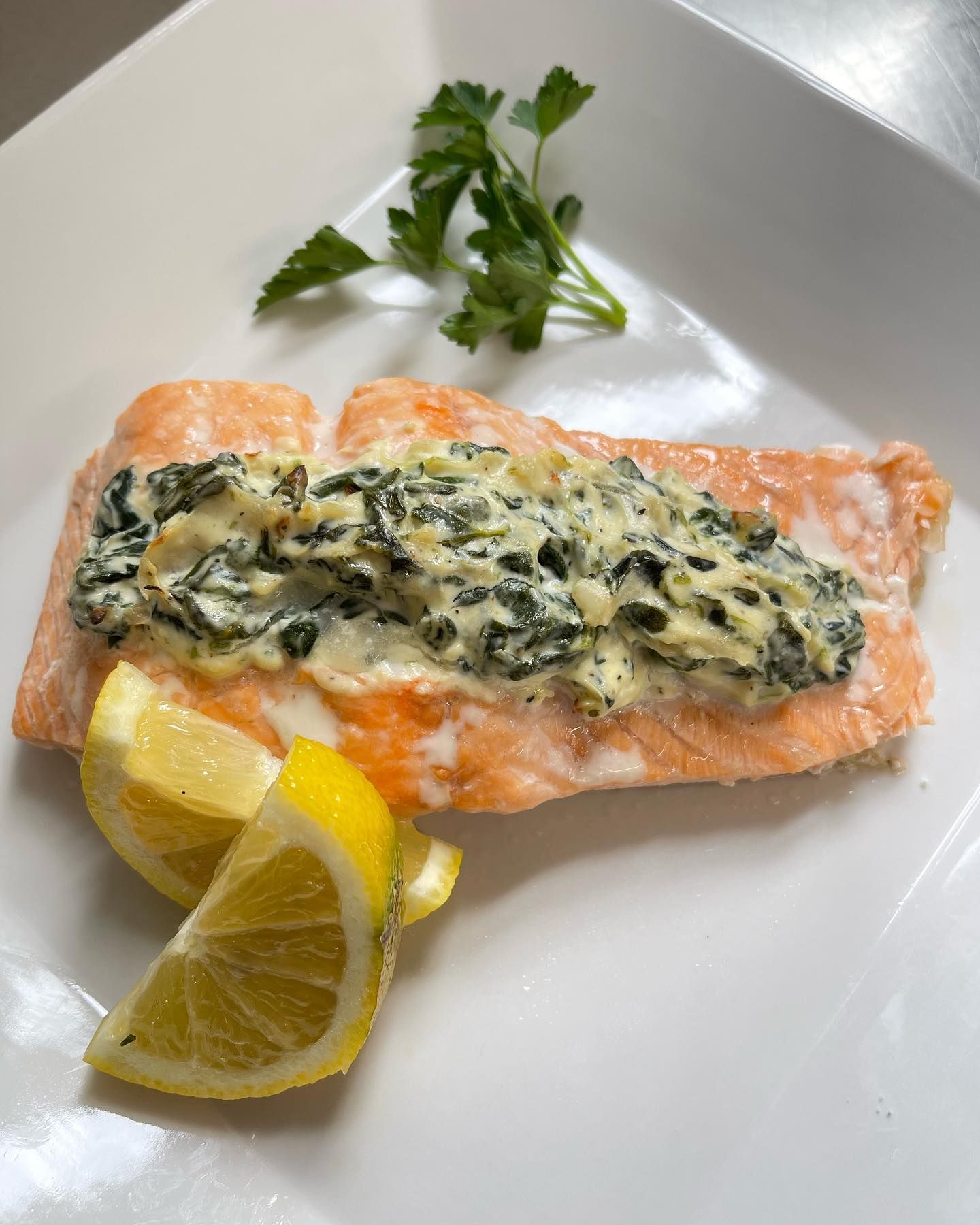 Salmon fillet topped with spinach and cream sauce, served with lemon slices and parsley garnish on a white plate.