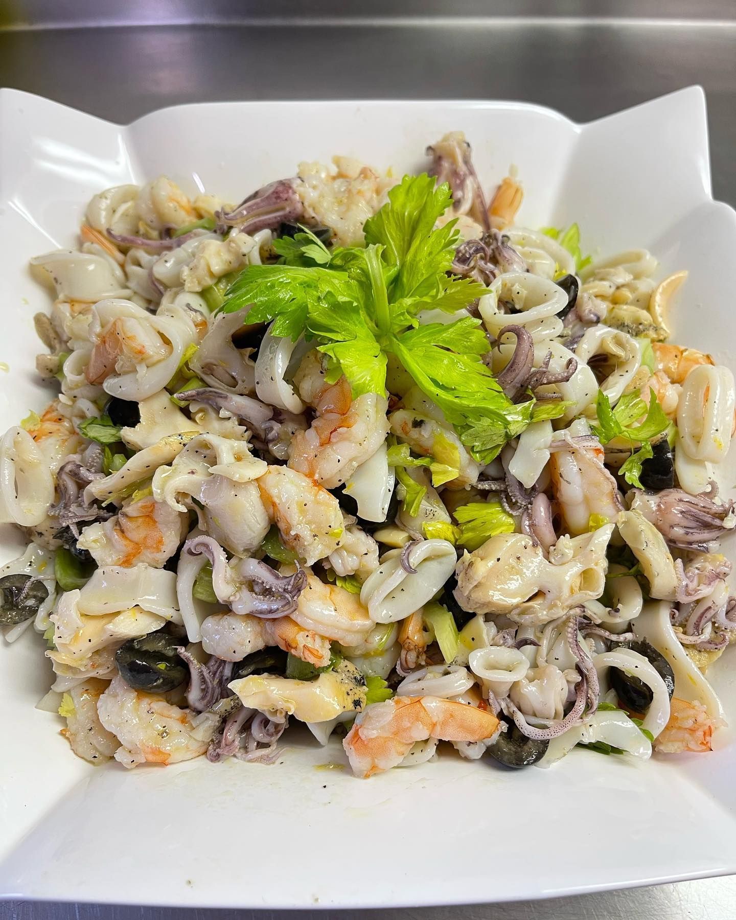 Seafood salad with shrimp, squid, and olives, garnished with celery, in a white dish.