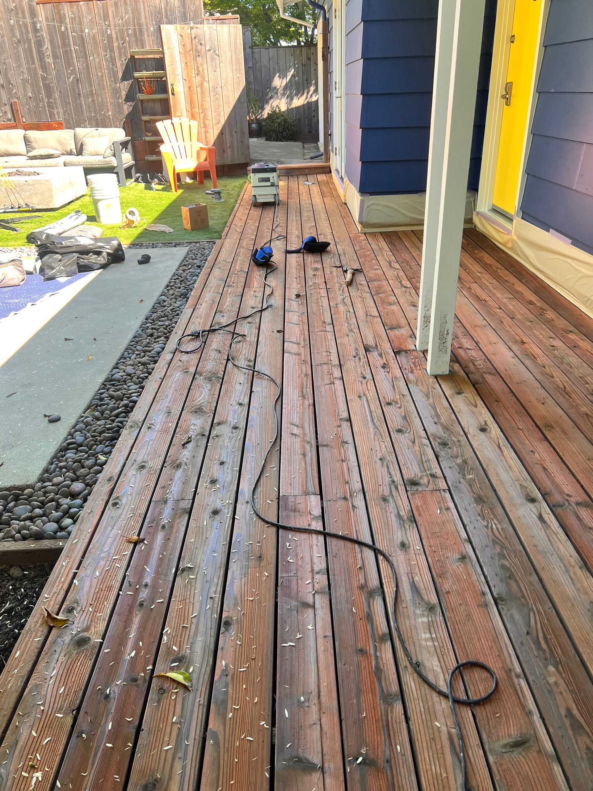 Wooden deck under renovation with scattered tools, cords, and fresh stain beside a blue house.