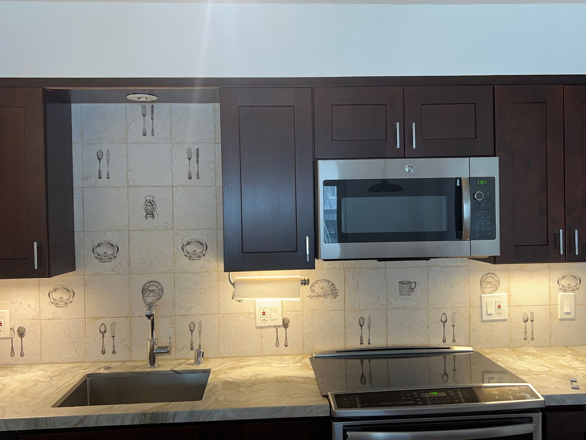 Modern kitchen with dark cabinets, granite countertop, tiled backsplash, sink, and stainless-steel microwave over stove