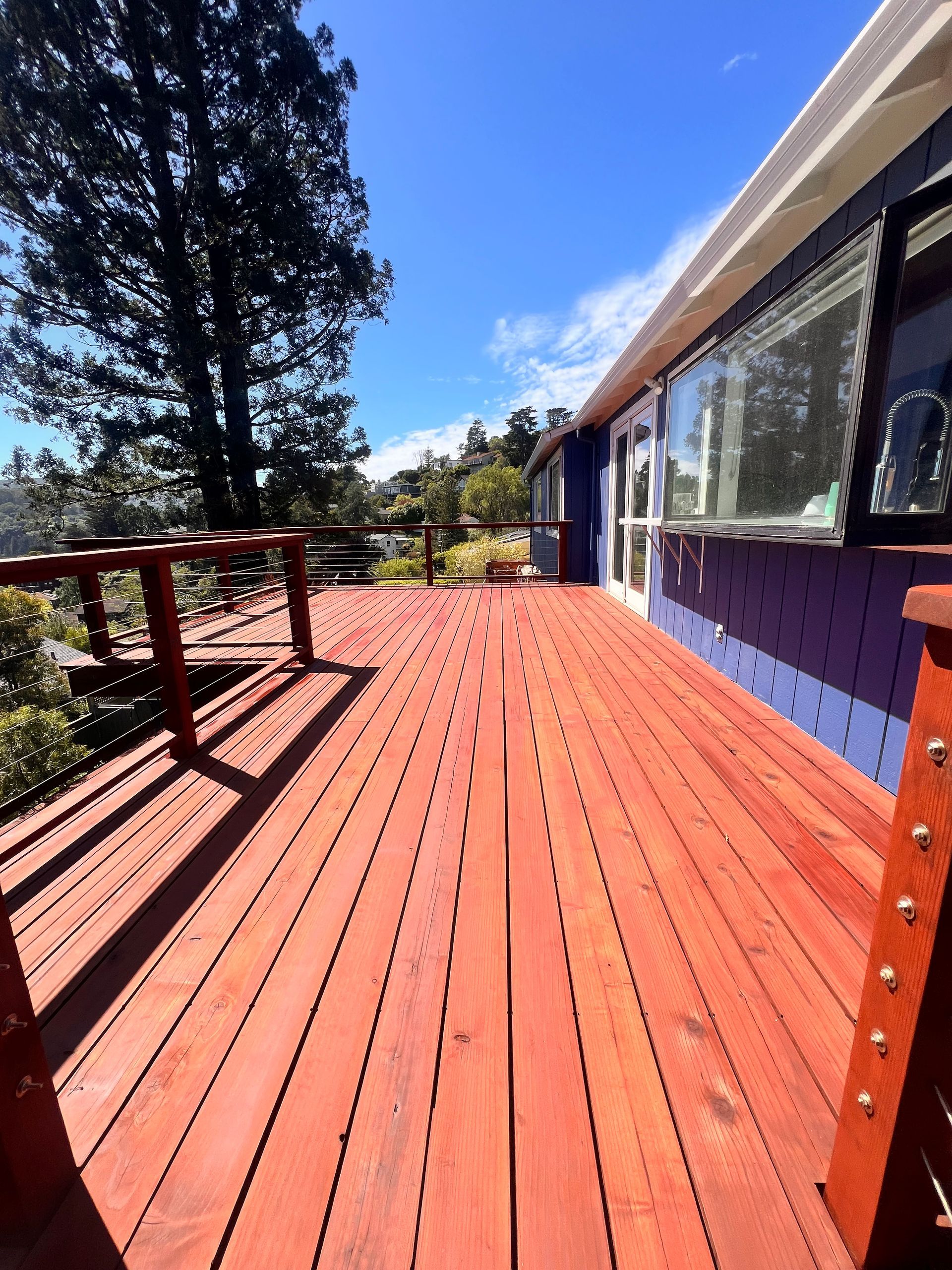 Red wooden deck beside a house with windows, under a bright blue sky and tall trees.