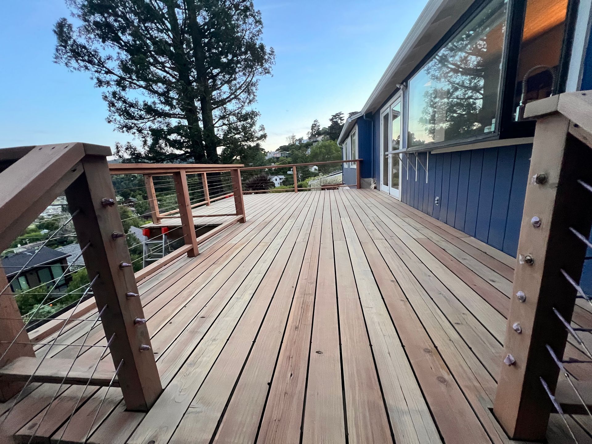 Wide wooden deck beside a blue house, with railings and a tree-lined view beyond.