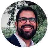 Hombre barbudo sonriente con gafas, en un primer plano circular, vistiendo una chaqueta oscura al aire libre.
