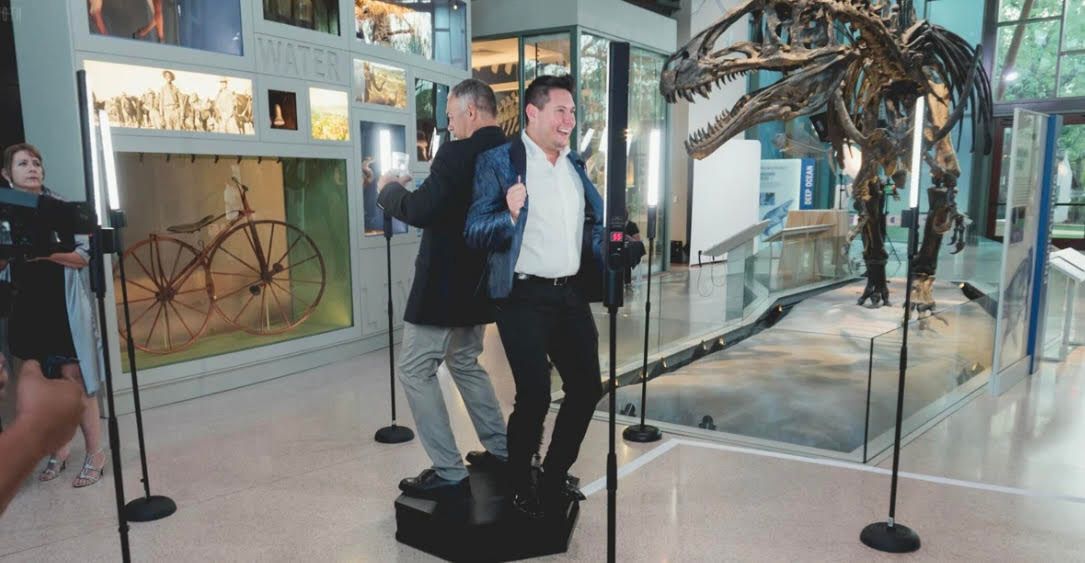 Two men are standing in front of a dinosaur skeleton in a museum.