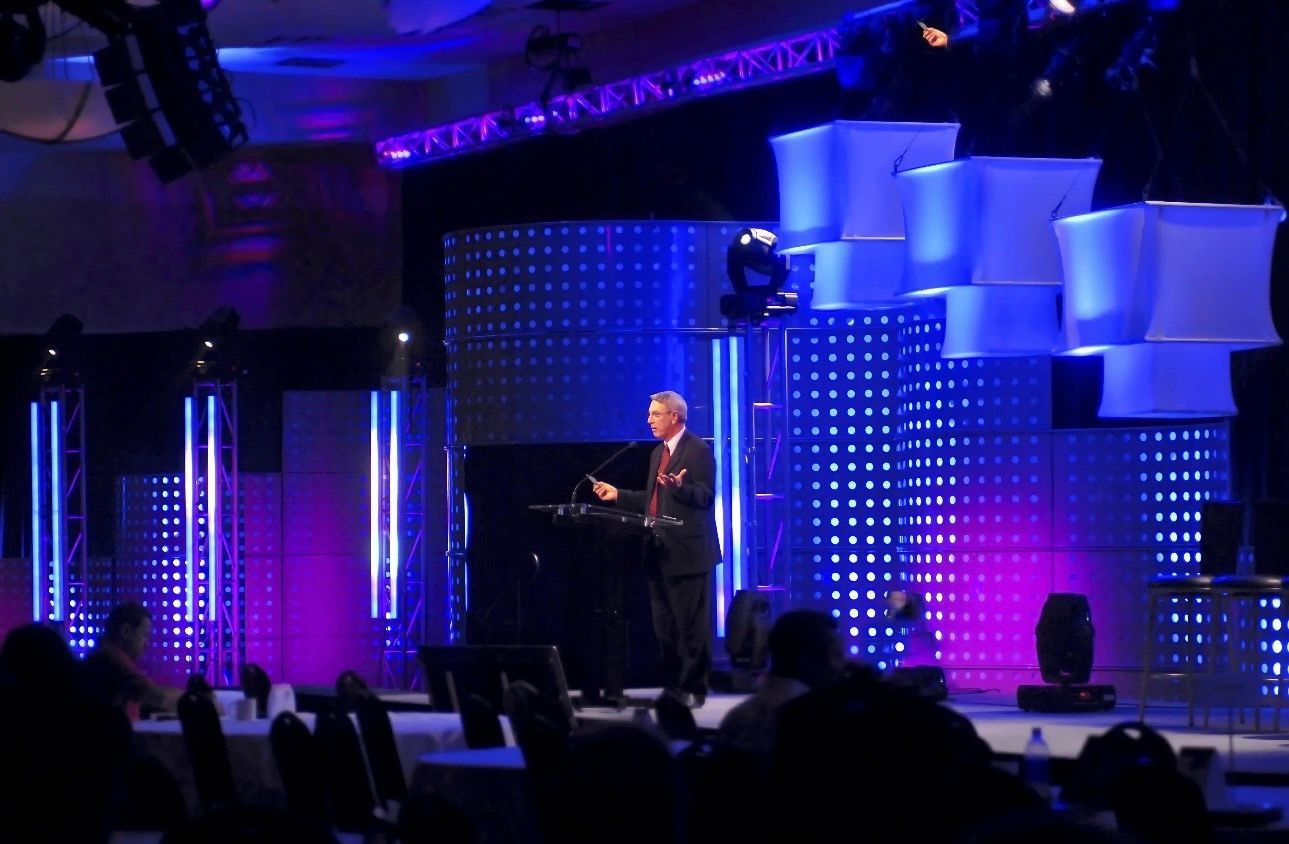 Speaker at podium on a purple-lit stage with geometric blue backdrop and audience silhouettes