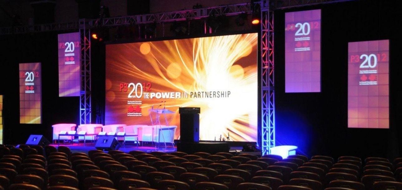 Conference stage with large central screen, purple side banners, and rows of empty seats in a dark auditorium