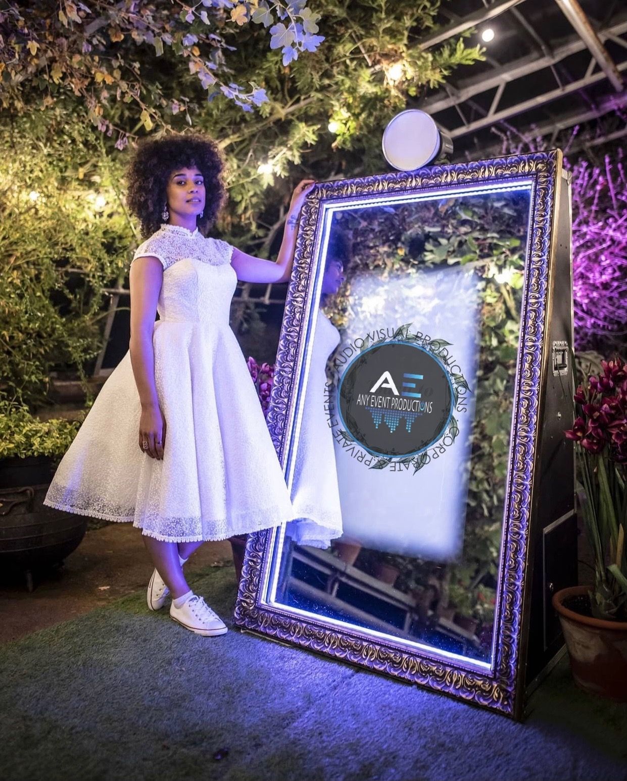 A woman in a white dress is standing next to a large mirror.