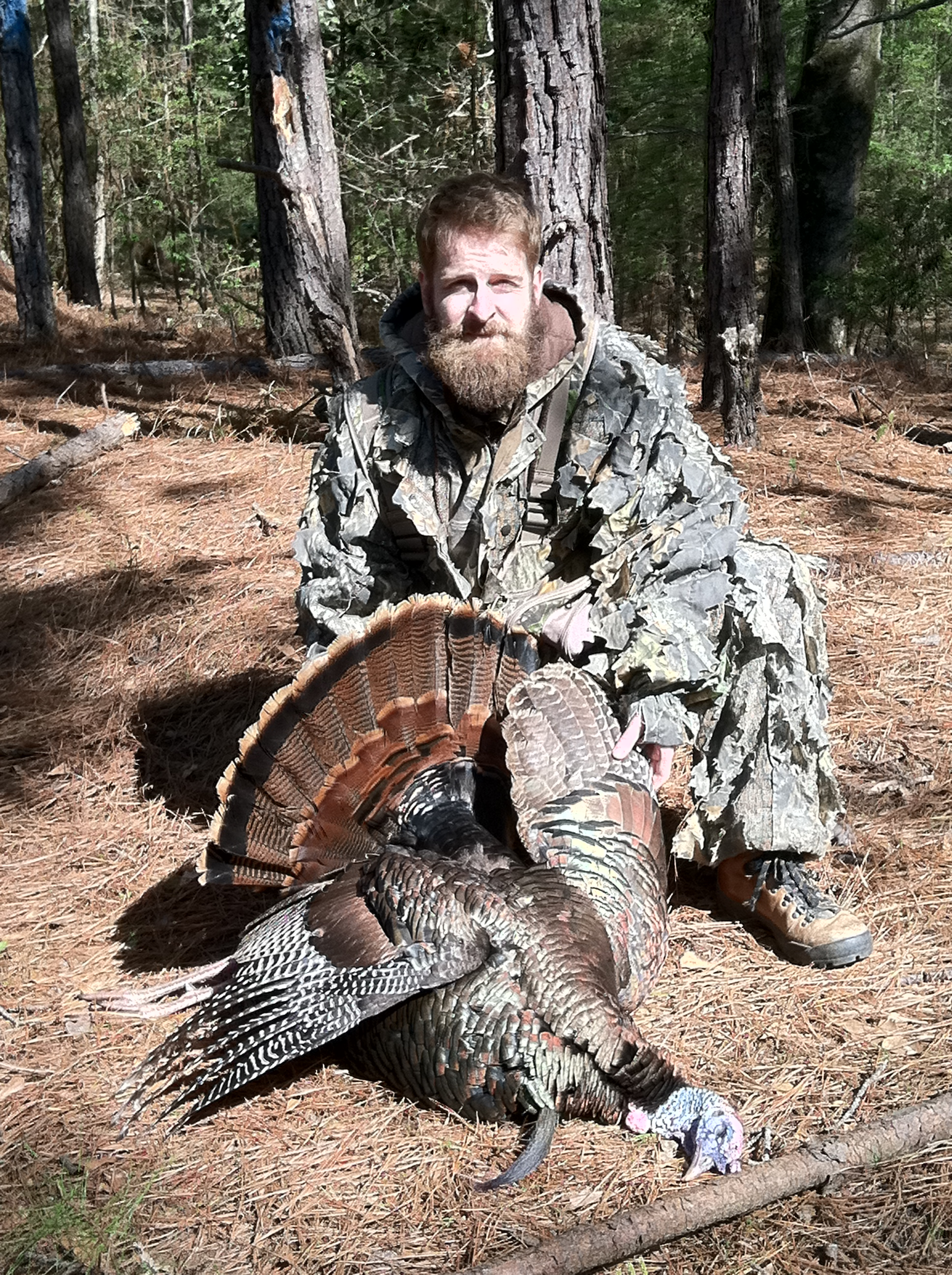 Man in camouflage kneeling next to a harvested wild turkey in a forest. He has a long beard and is smiling.