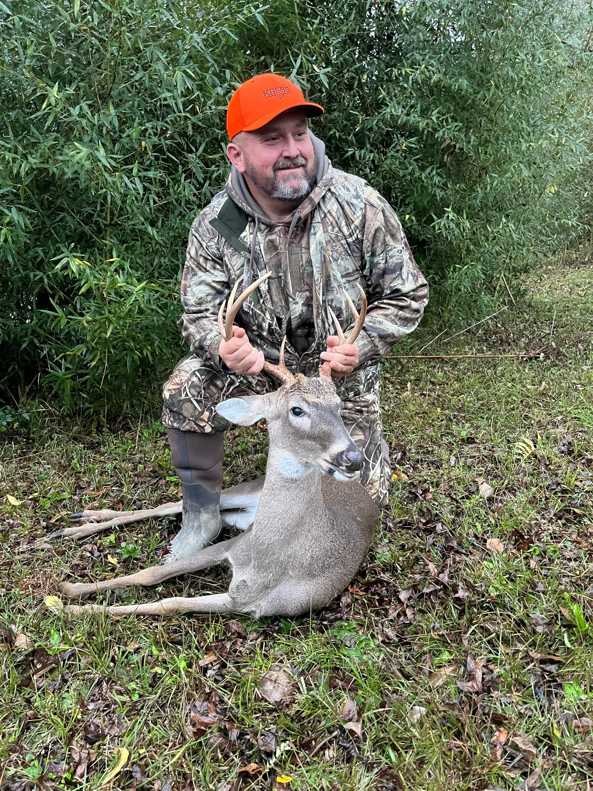 Hunter kneeling with a harvested buck, outdoors. He wears camo and an orange hat, smiling, holding the deer's antlers.