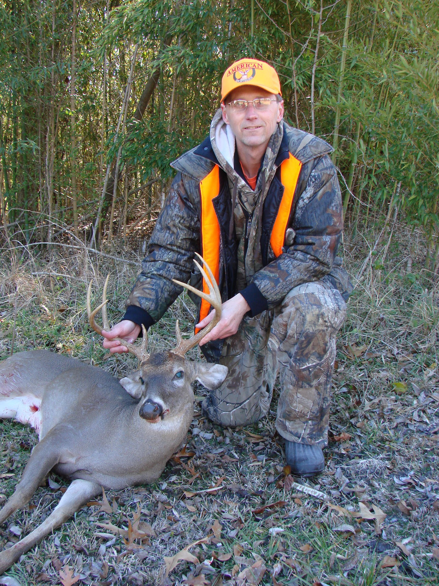 Hunter kneeling beside a harvested deer in a wooded area. The hunter wears camouflage and orange safety gear.