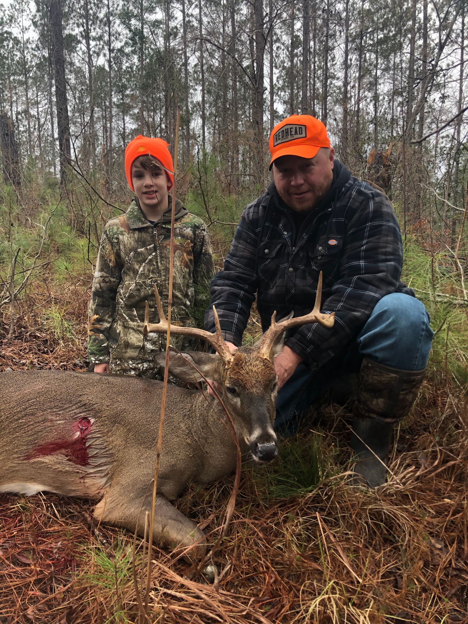 A man and a boy pose with a dead deer in a forest. Both wear orange hats. The deer has antlers.
