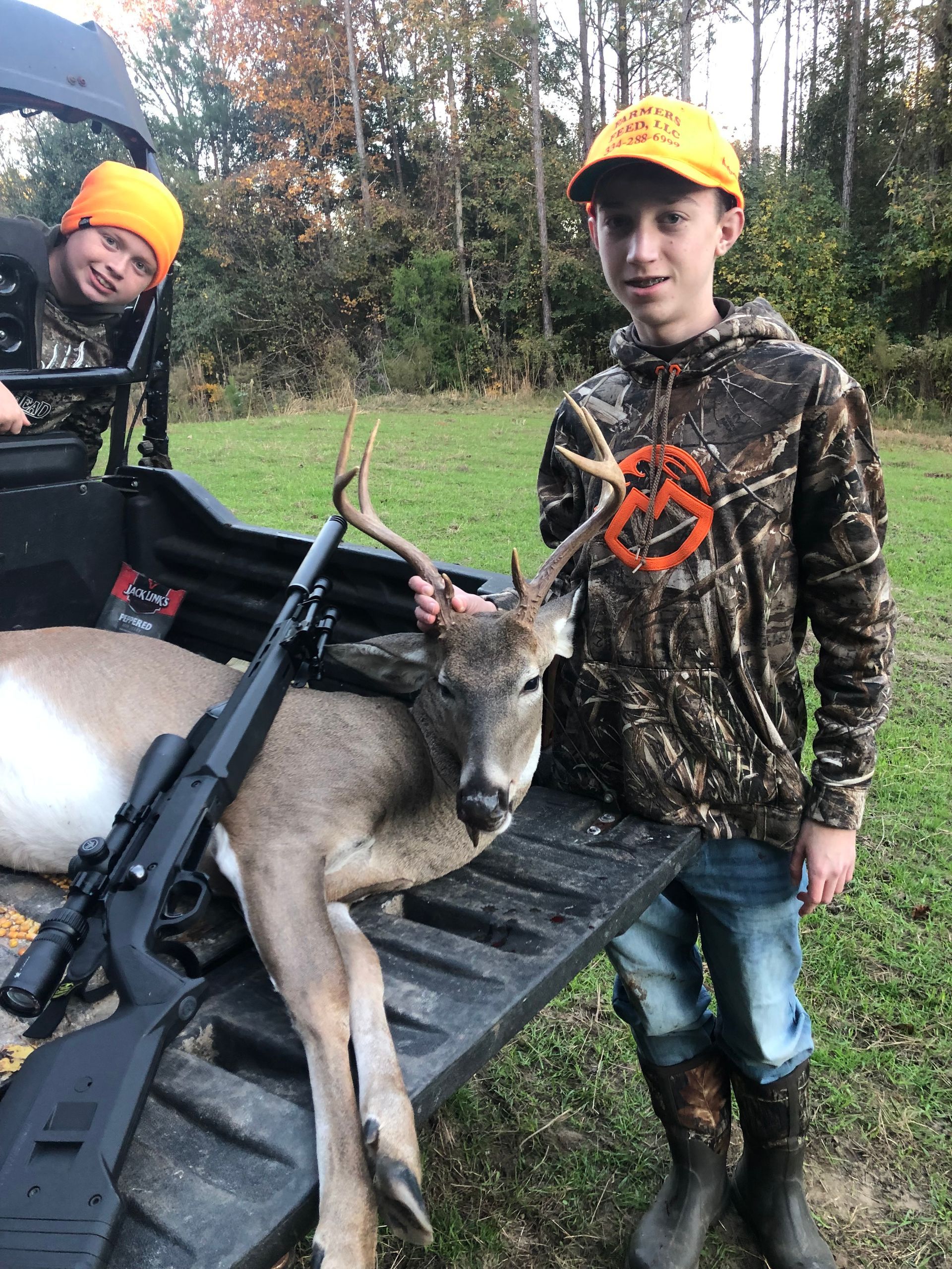 Two young boys in hunting gear pose with a deer in the back of a vehicle. One boy holds the deer's antlers, while a rifle rests nearby.