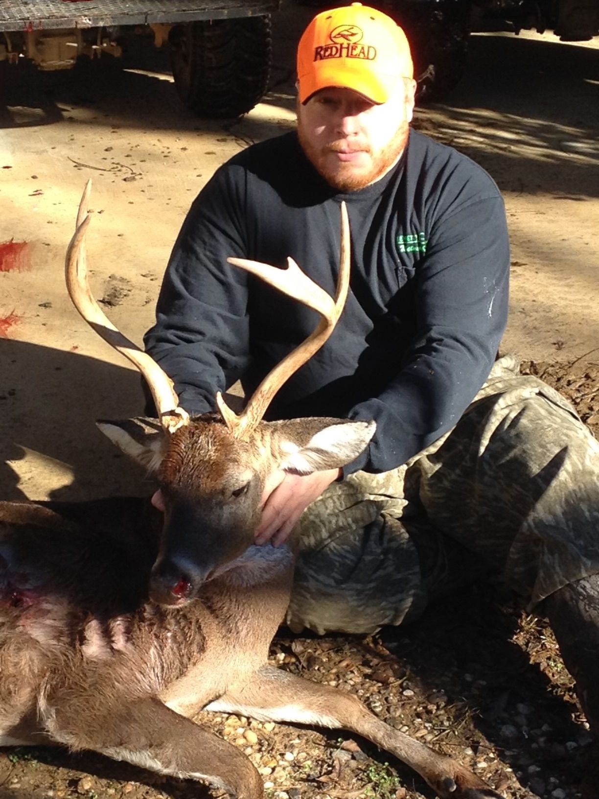 Hunter in orange hat and black shirt kneels next to a dead buck with antlers. Bloodstains present.