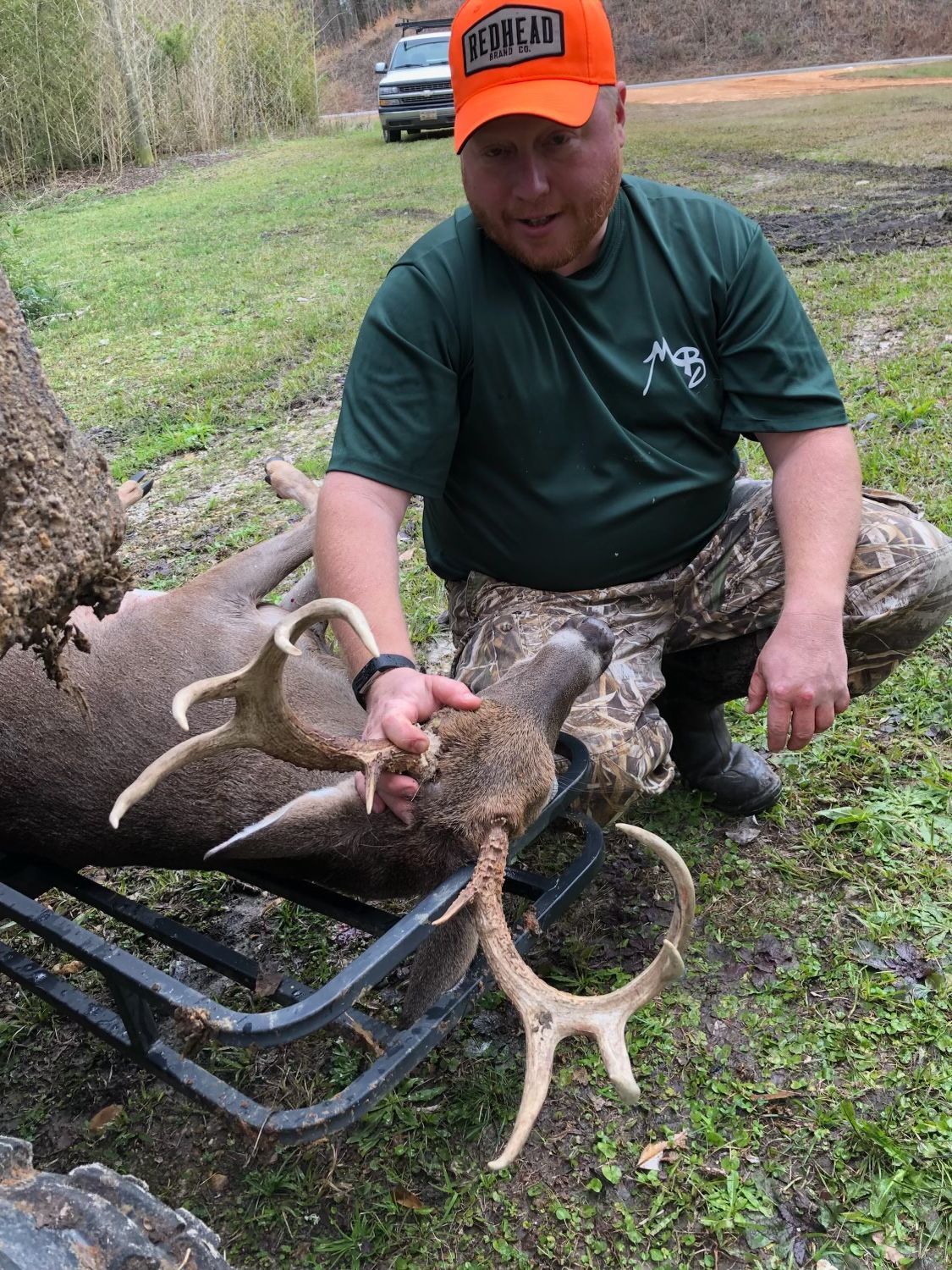 A man kneels beside a harvested deer with large antlers, wearing a blaze orange cap and camo pants outdoors on a cloudy day.