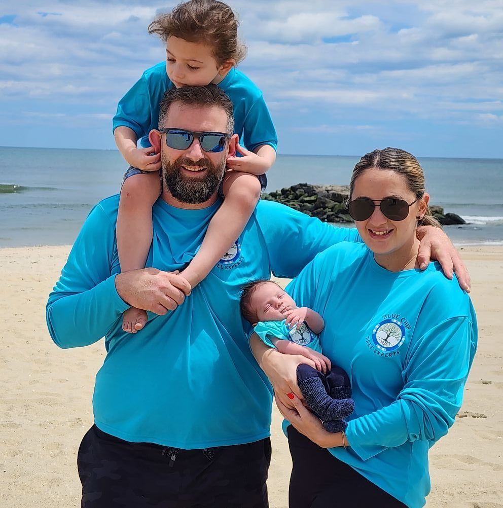 Family of four on a beach; father with child on shoulders, mother holding baby, all wearing blue shirts, sunny sky.