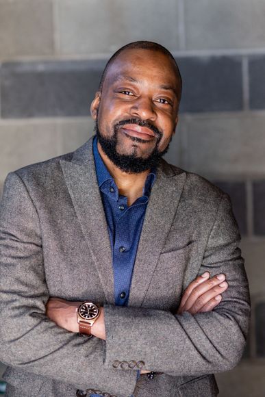 Smiling man in a gray blazer and blue shirt, standing with arms crossed against a tiled wall.