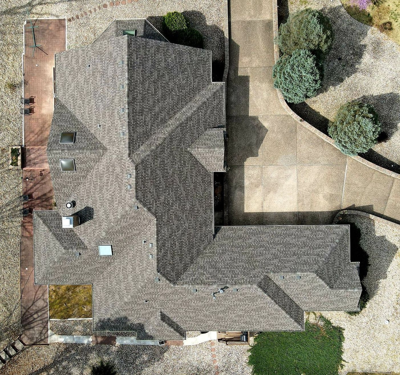 Aerial view of a house roof with gray shingles, surrounding patio, and trees.