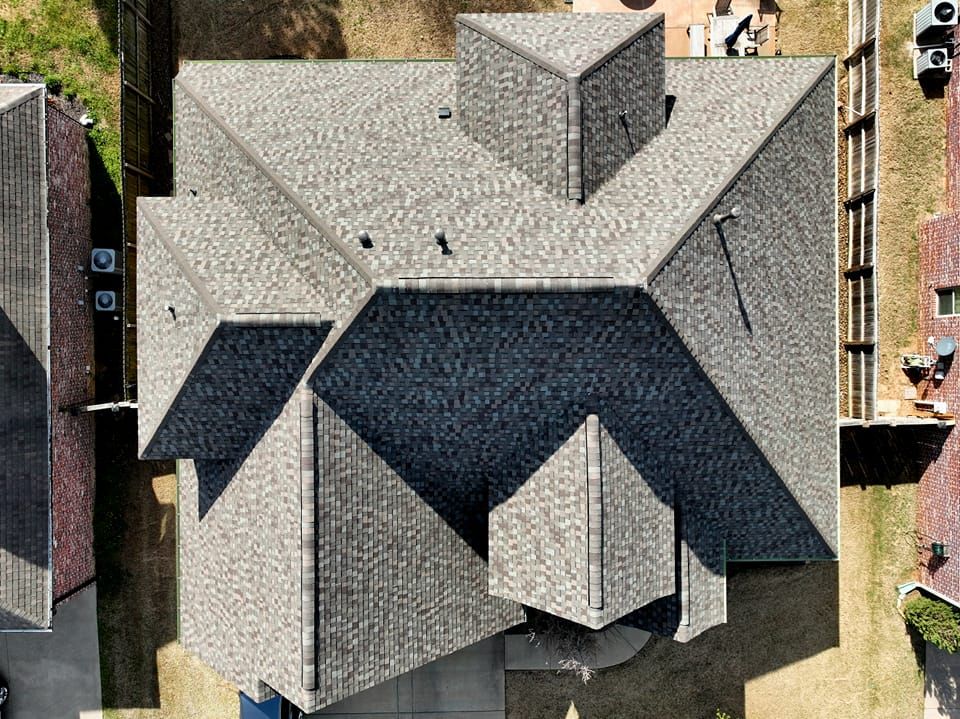 Aerial view of a gray-shingled house roof with multiple gables and a central chimney-like structure.