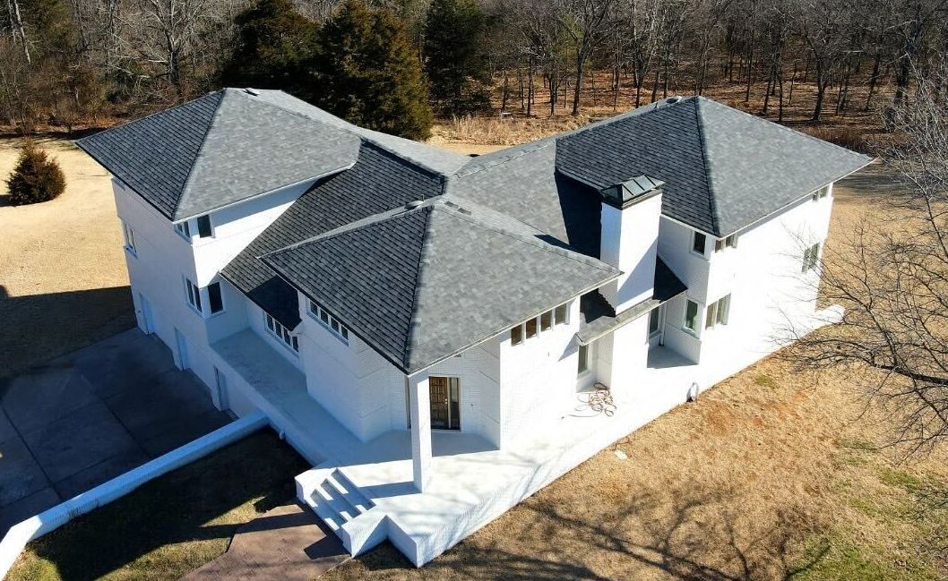 Aerial view of a white house with a gray roof surrounded by trees and dry grass.