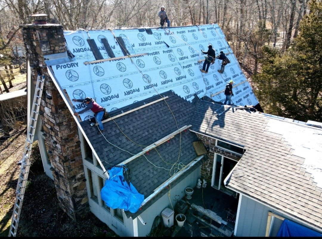 Workers installing roof underlayment on a house roof, with blue tarp and trees in the background