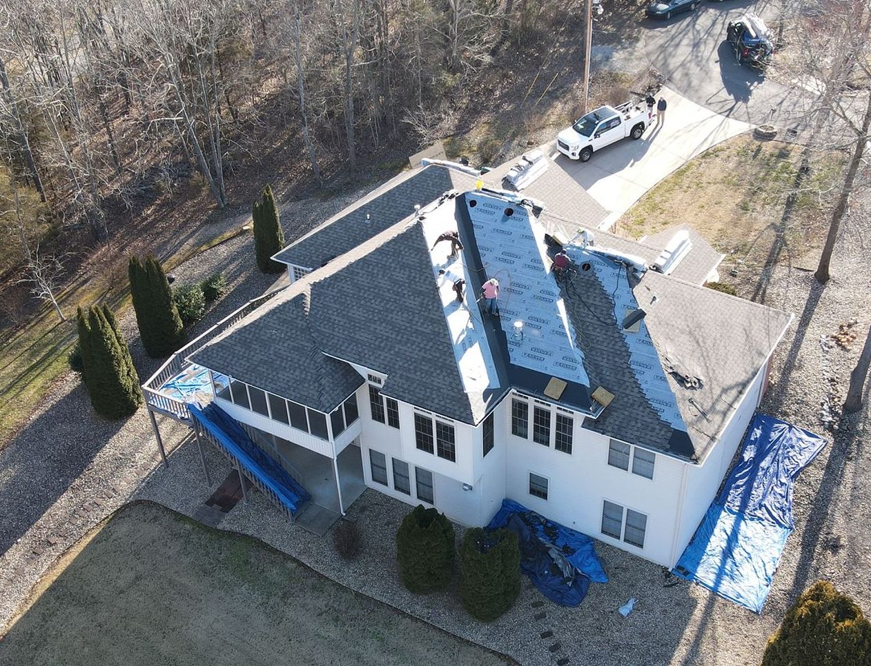 Aerial view of a white house with a dark roof, driveway, and surrounding bare trees