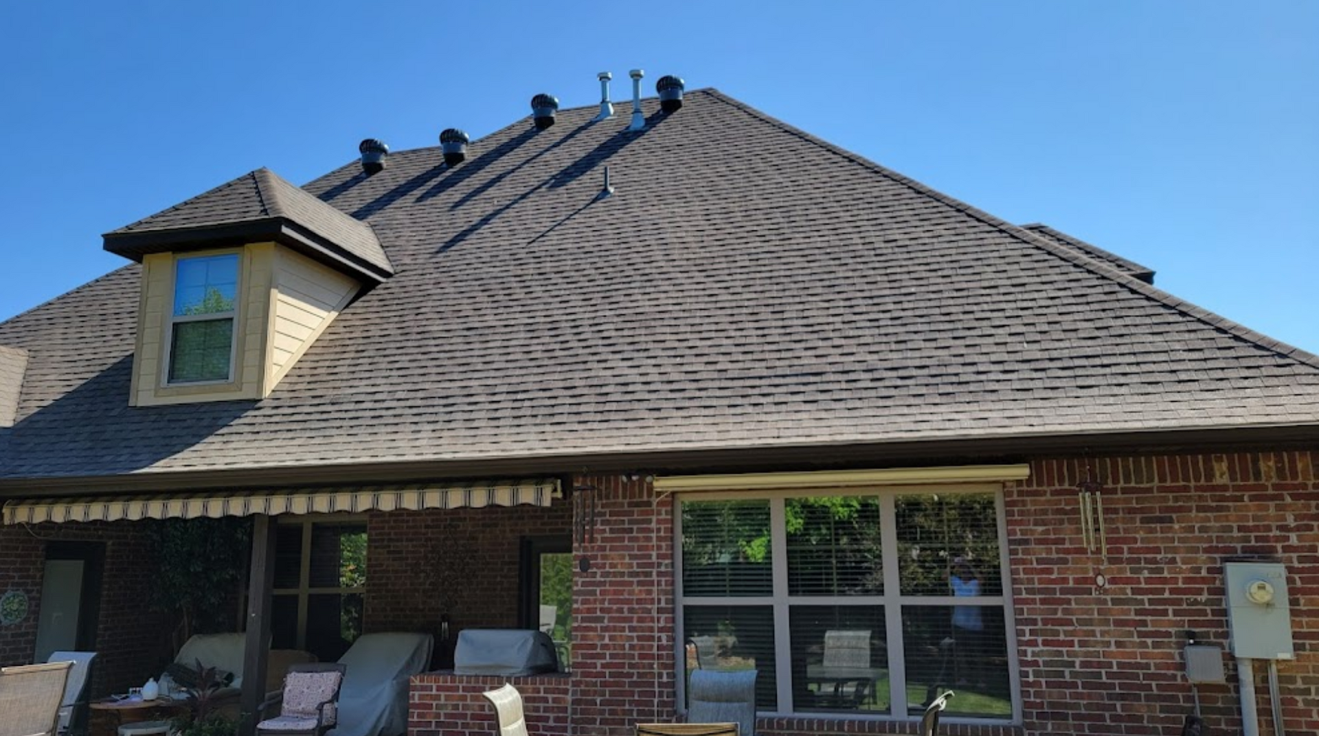Brick house with large shingled roof, dormer window, and backyard patio chairs under a clear blue sky