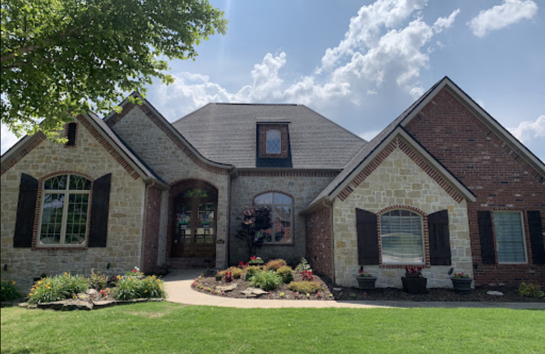 Brick suburban house with arched entry, dark shutters, manicured lawn, and flower beds under a blue sky
