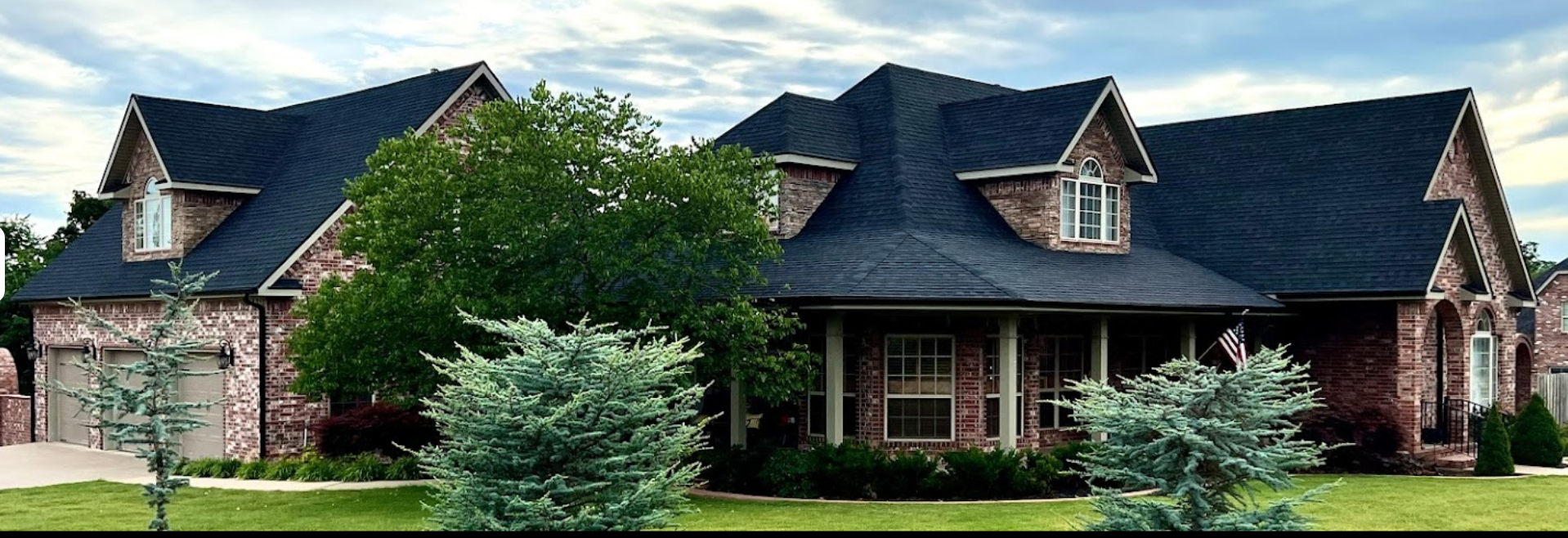 Brick suburban house with dark roof, front porch, and landscaped trees and shrubs