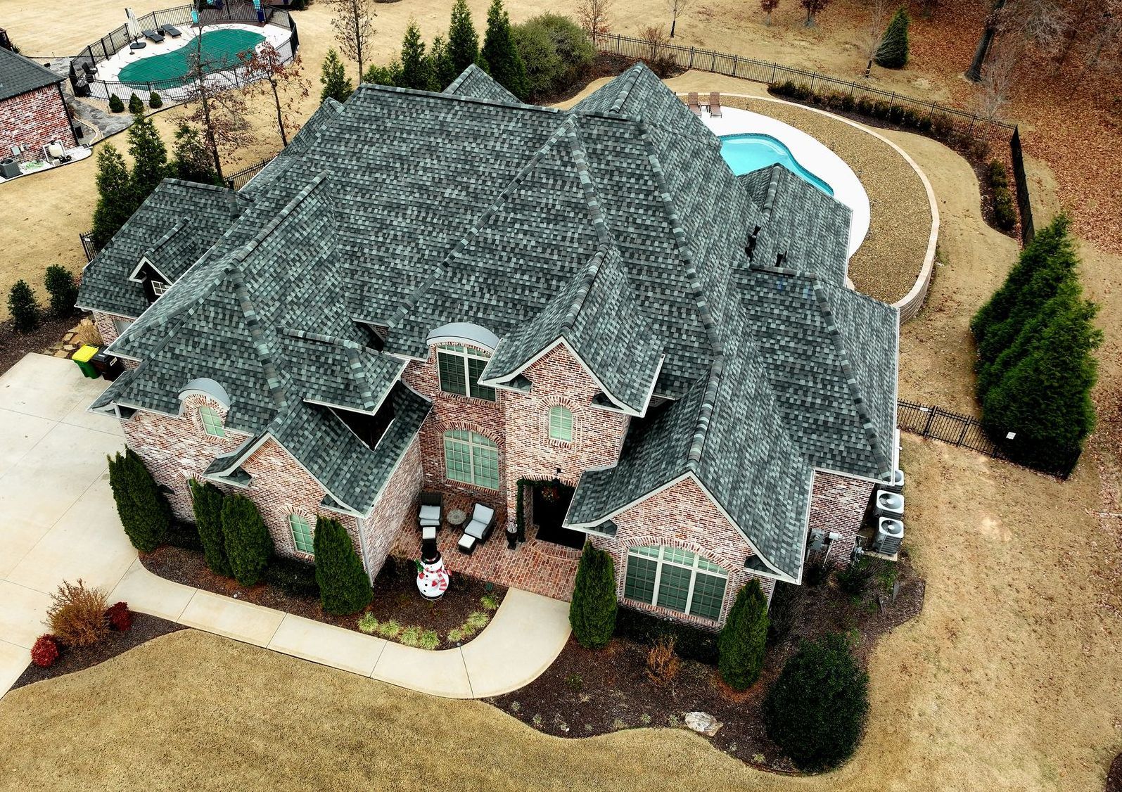 Aerial view of a large brick house with a gray roof, surrounded by tan lawn, trees, and a curved driveway.