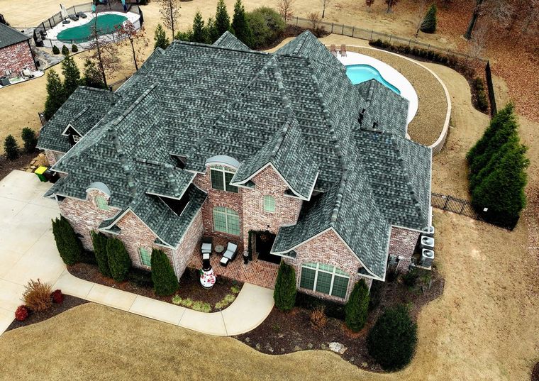 Aerial view of a large brick house with a gray roof, surrounded by tan lawn, trees, and a curved driveway.